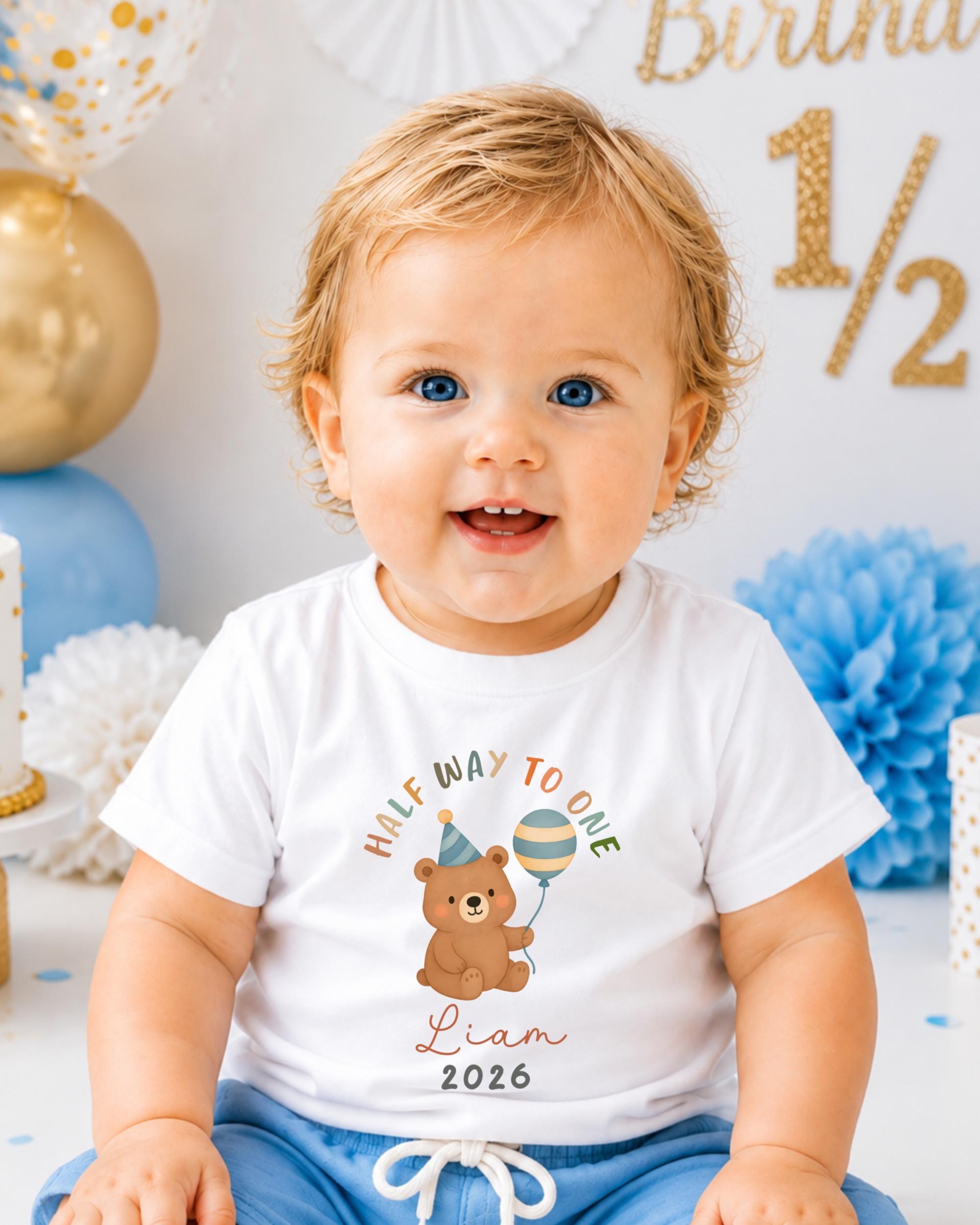 A smiling baby boy wearing a white t-shirt with a cartoon bear and text, sitting in front of a birthday party decoration with gold and blue balloons.