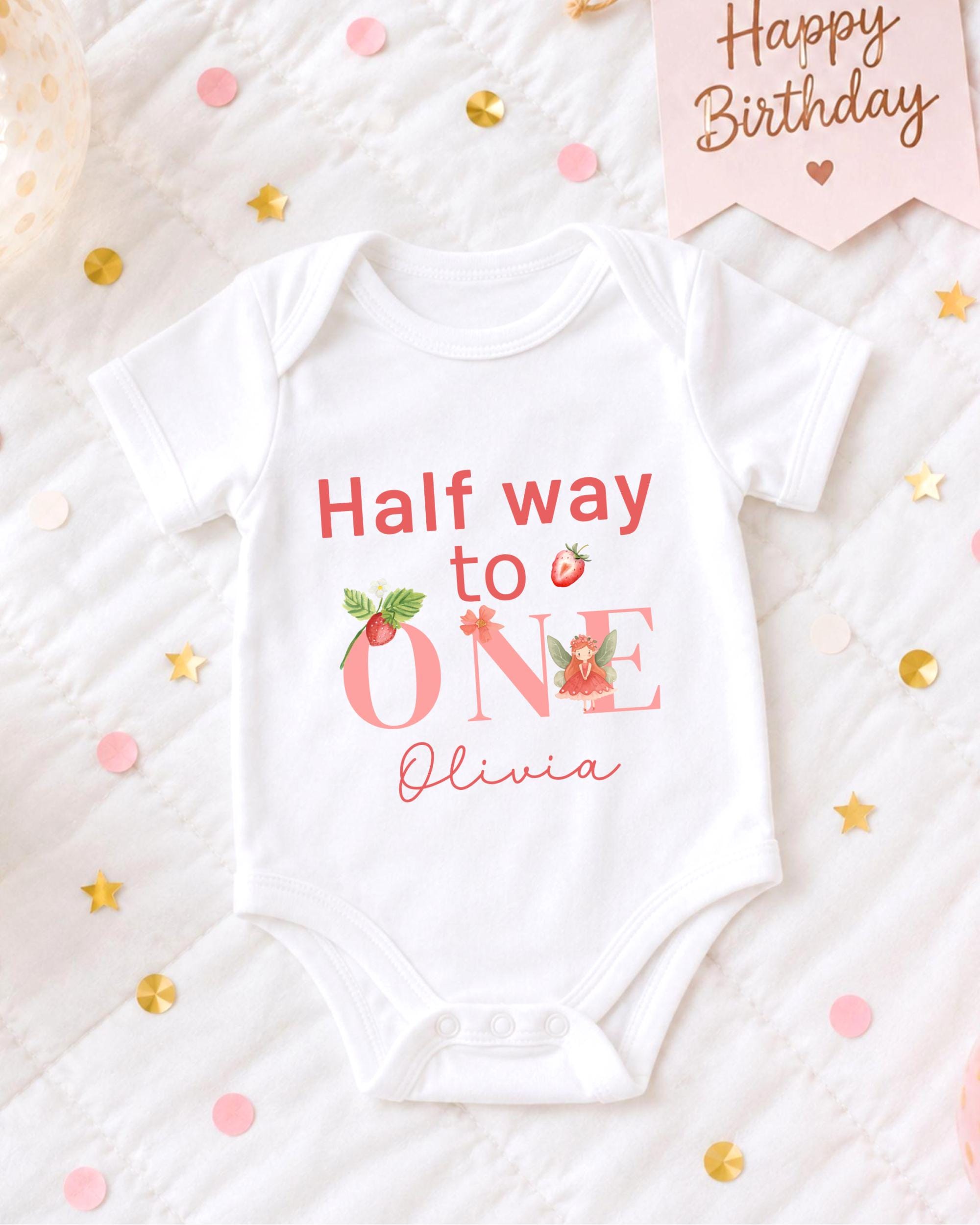A white onesie with the text "Half way to one" printed on it, along with the name "Olivia" below it. The onesie is placed on a bed with confetti and a birthday card.