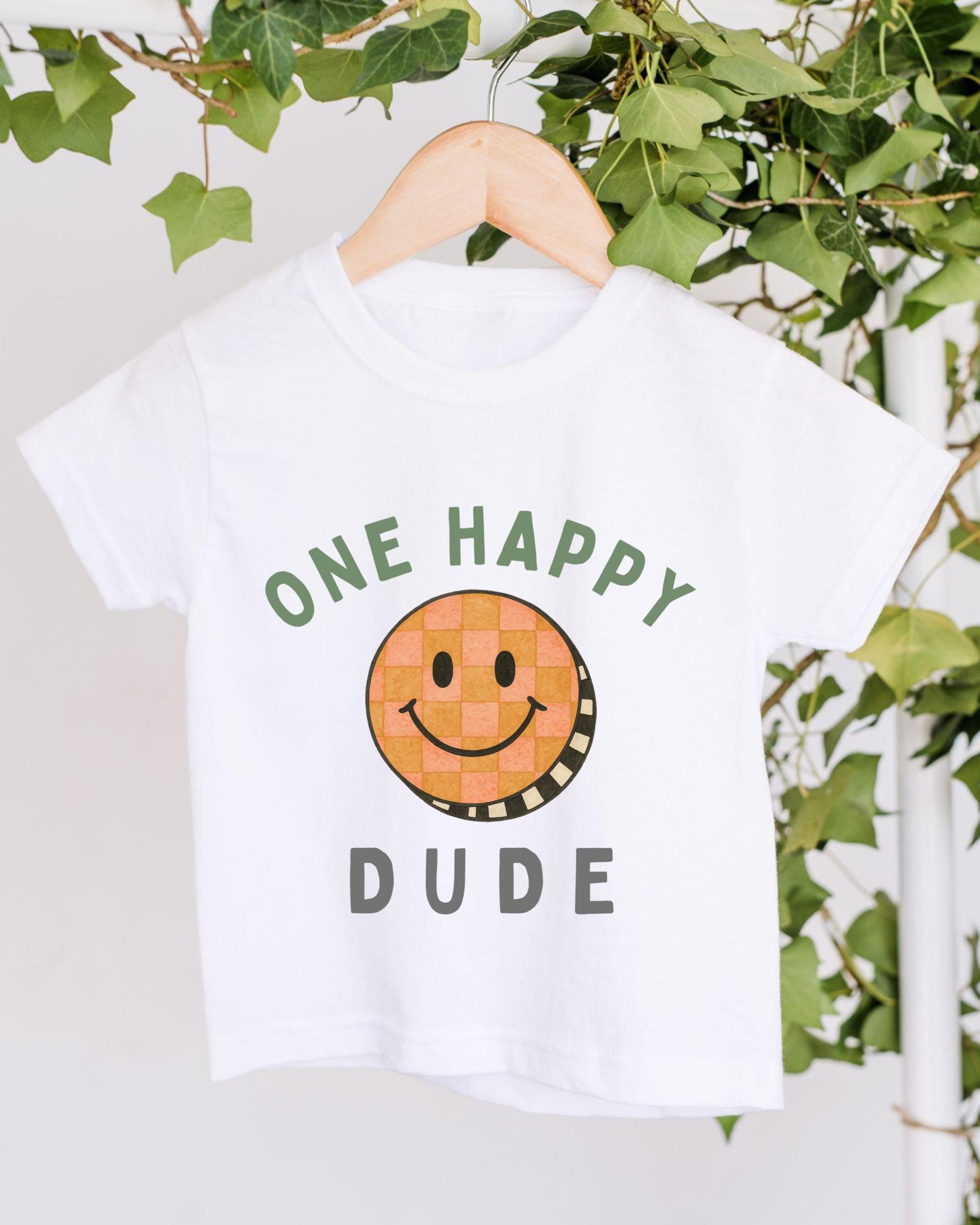 A white t-shirt with a cheerful smiley face and the text "ONE HAPPY DUDE" printed on it, hanging on a wooden hanger.