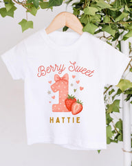 A white t-shirt with a personalized design hanging on a wooden hanger. The design features a pink polka dot number one, a strawberry, and the text "Berry Sweet" and "HATTIE".