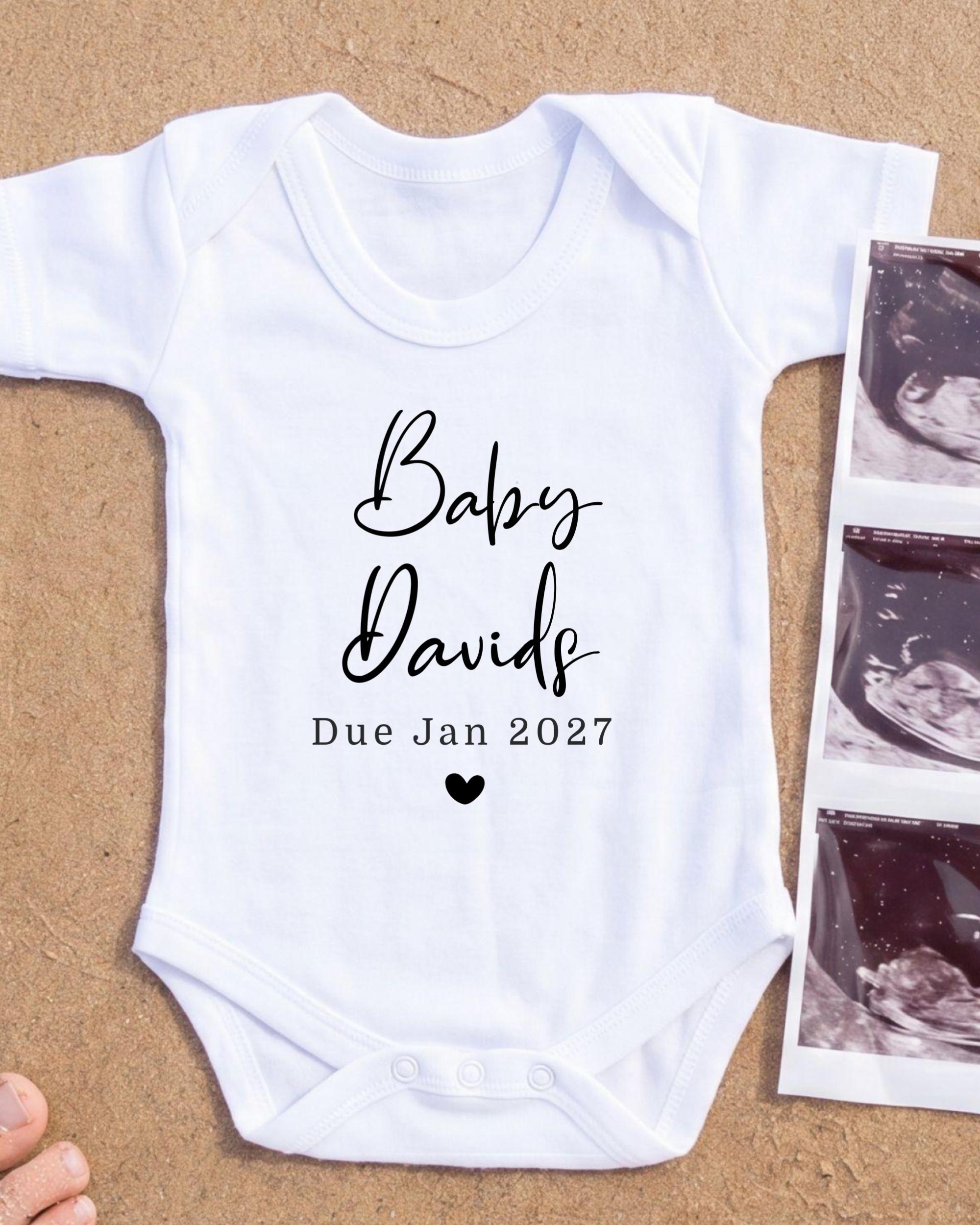 A white baby onesie with the text "Baby Davids Due Jan 2027" printed on it, along with a heart symbol. The onesie is placed next to a photograph of an ultrasound image.