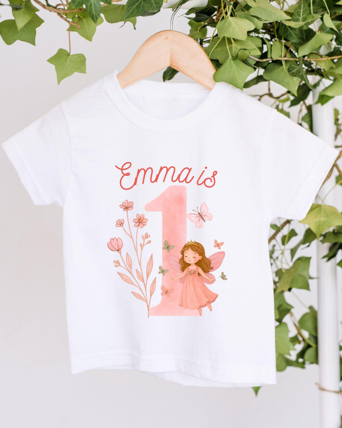 A white t-shirt with a pink number one and a fairy design, along with the text "Emma is 1" printed on it.