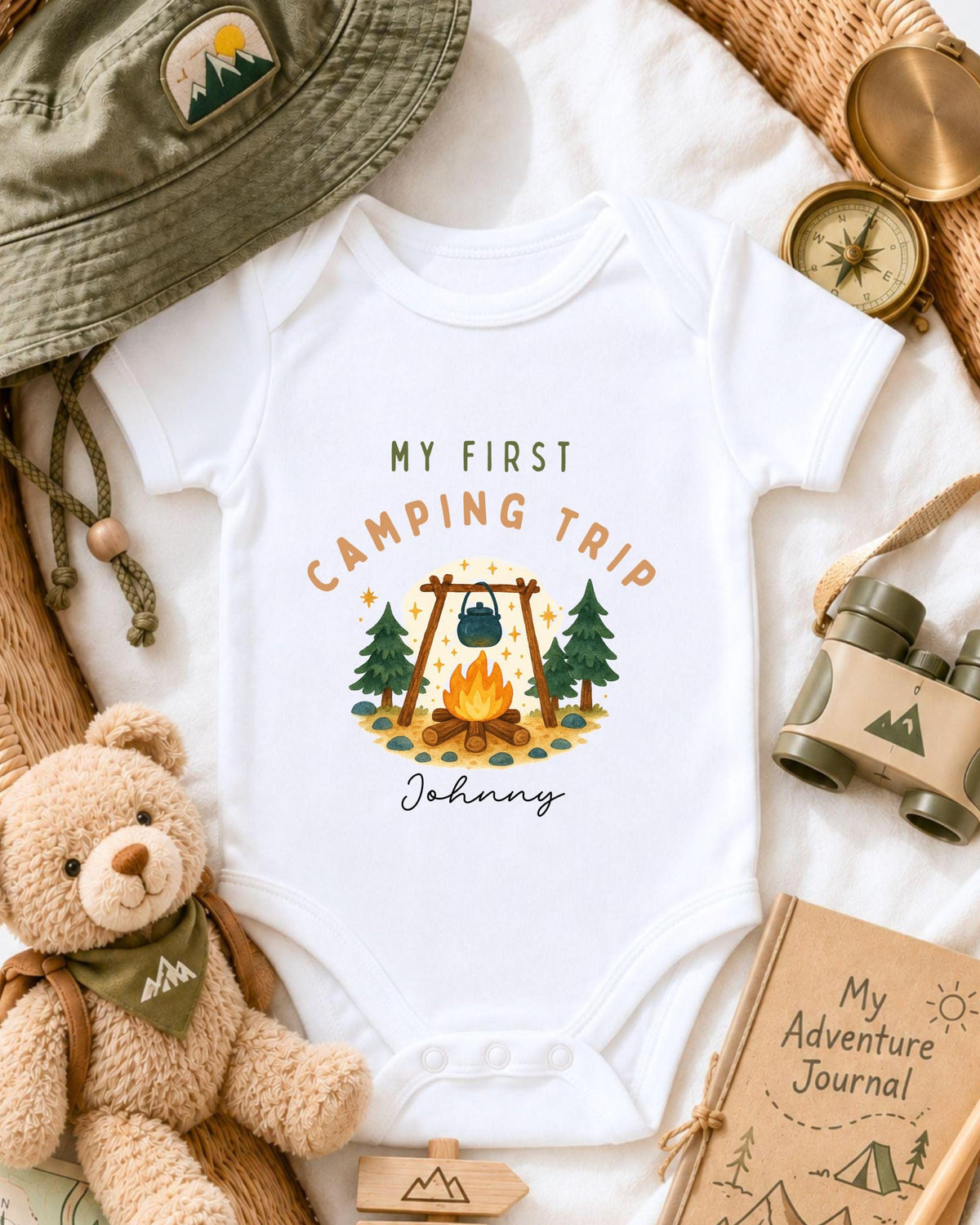 a baby onesie with a camping-themed design, surrounded by camping-related items such as a hat, compass, binoculars, and a teddy bear.