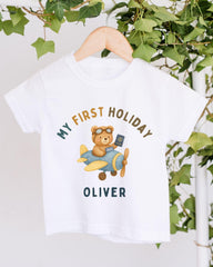 A white t-shirt with a graphic design of a teddy bear wearing sunglasses and holding a suitcase, with the text "My first holiday Oliver" printed on it.