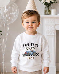 A young boy wearing a white sweatshirt with a cartoon race car and the text "Two Fast Jack" is standing in front of a fireplace.