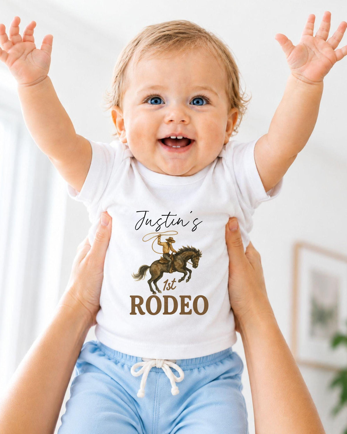A smiling baby wearing a white t-shirt with a cowboy graphic and the text " Justin's 1st Rodeo" is being held up by someone's arms.