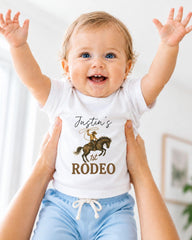 A smiling baby wearing a white t-shirt with a cowboy graphic and the text " Justin's 1st Rodeo" is being held up by someone's arms.