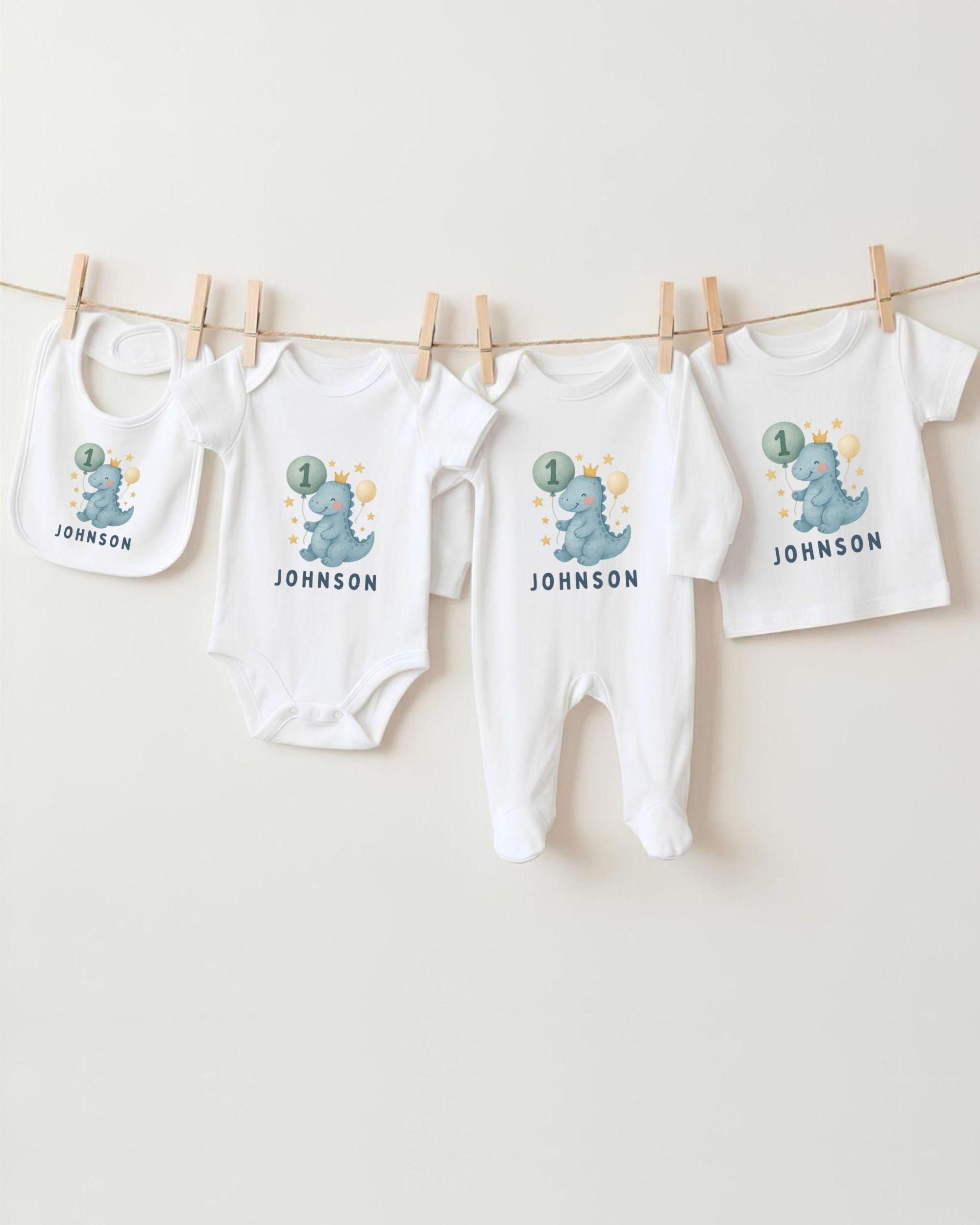 A collection of white baby clothes, including onesies and bibs, with dinosaur designs and the name "Johnson" printed on them.