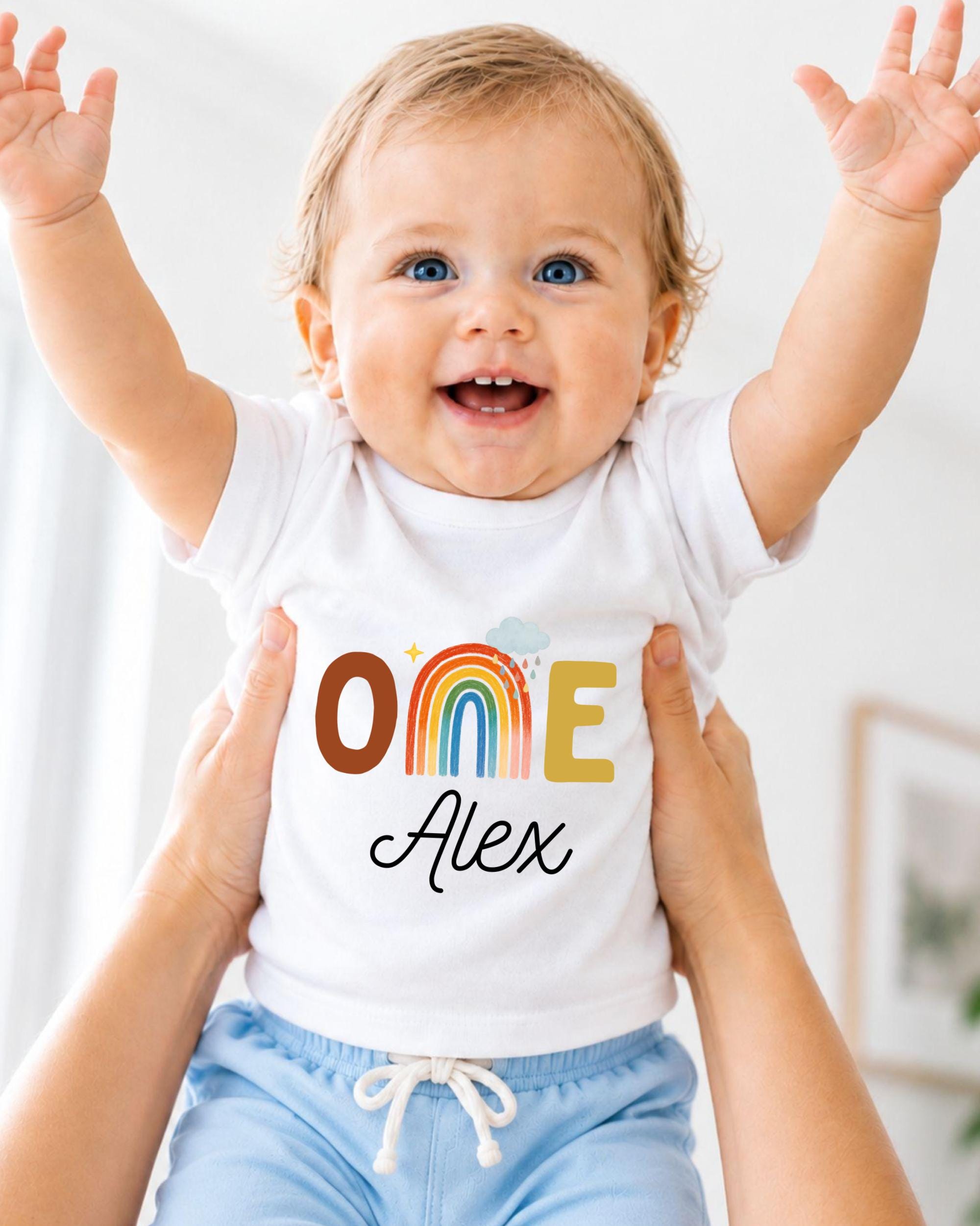 A smiling baby wearing a white t-shirt with the name "Olive" and the name "Alex" printed on it, with their arms raised in the air.
