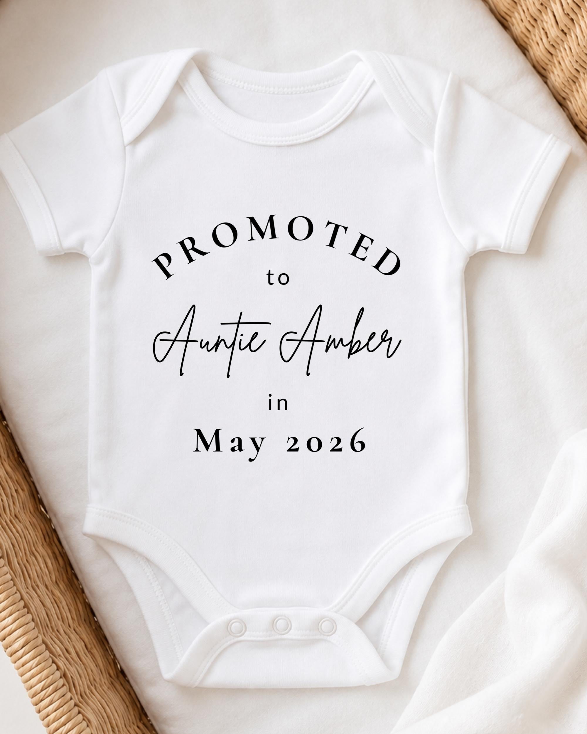 A white baby onesie with text printed on it, placed on a bed with a woven blanket.