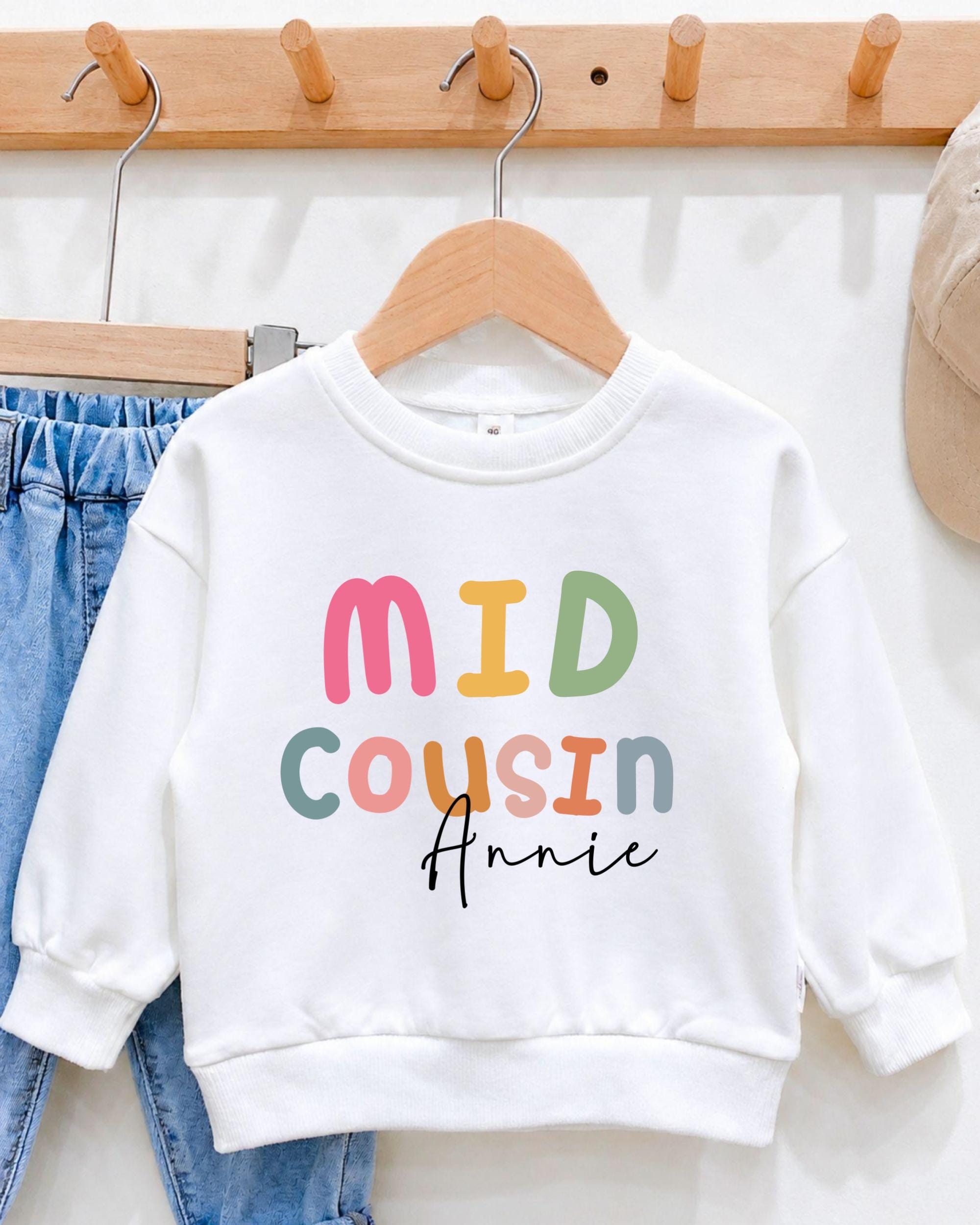 A white sweatshirt with the text "MID COUSIN Annie" printed on it, hanging on a wooden hanger.