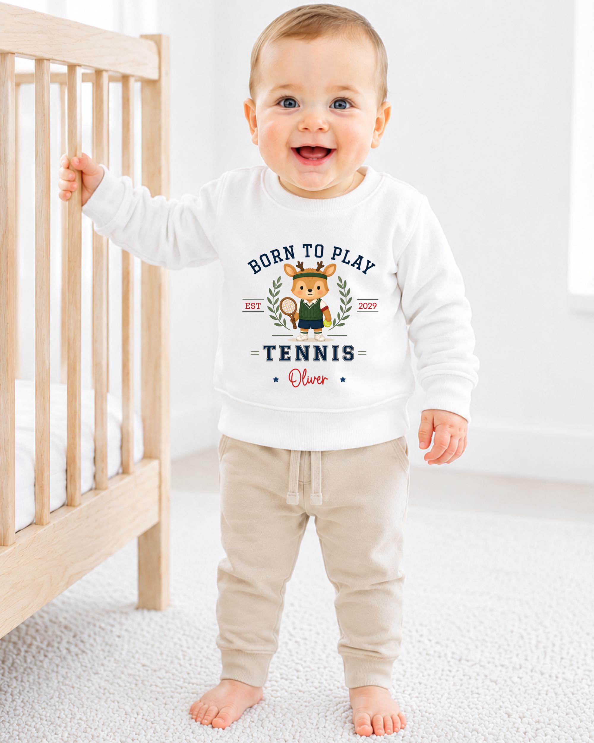 A smiling baby boy wearing a white sweatshirt with a tennis-themed design and text.