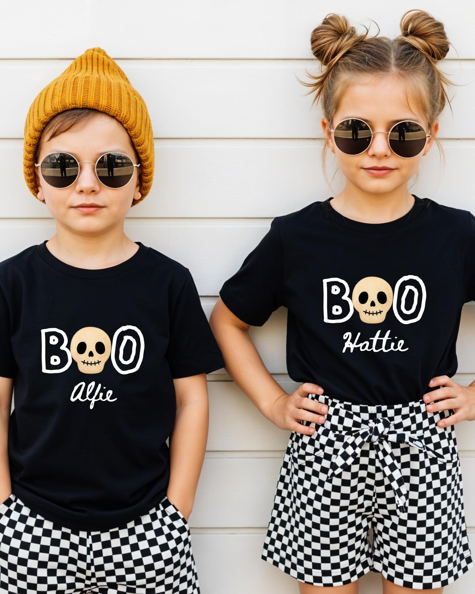 Two children wearing black t-shirts with the text "BOO" and "Alfie" printed on them, along with skull designs, stand against a white wall.