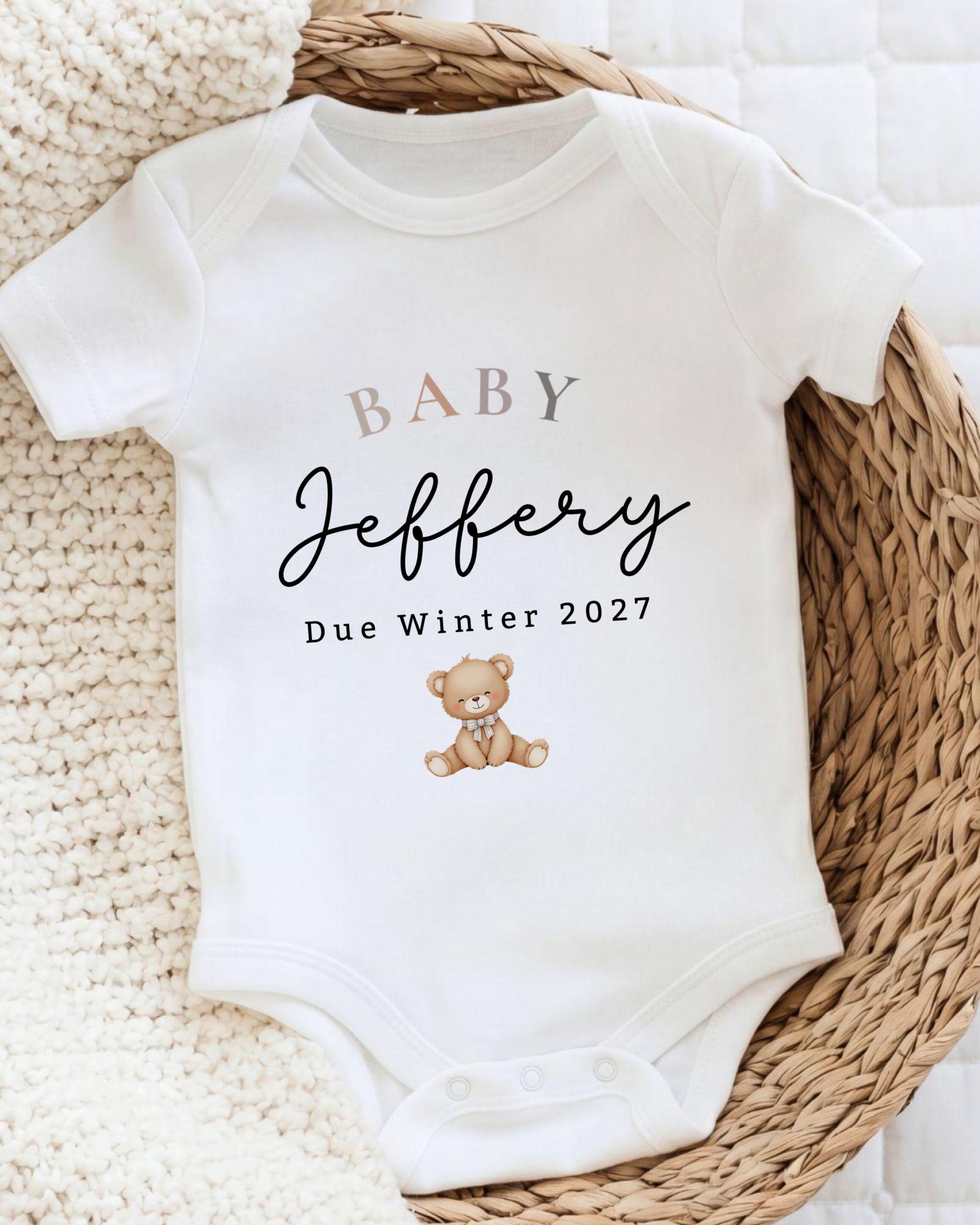 A white baby onesie with the text "BABY Jeffery Due Winter 2027" printed on it, along with a teddy bear image.