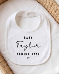 A white baby bib with the text "BABY Taylor COMING SOON" printed on it, placed on a white cloth.