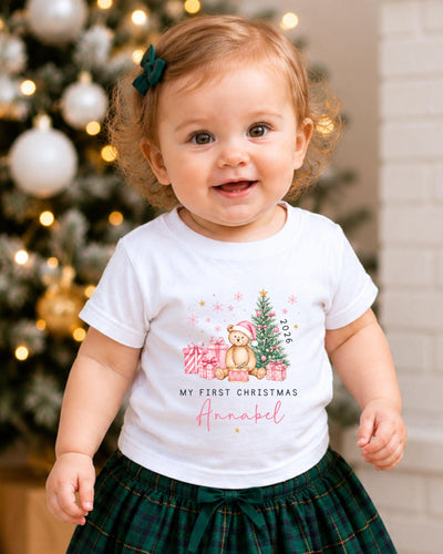 A smiling baby girl wearing a white t-shirt with a Christmas-themed design and a green bow in her hair, standing in front of a Christmas tree.
