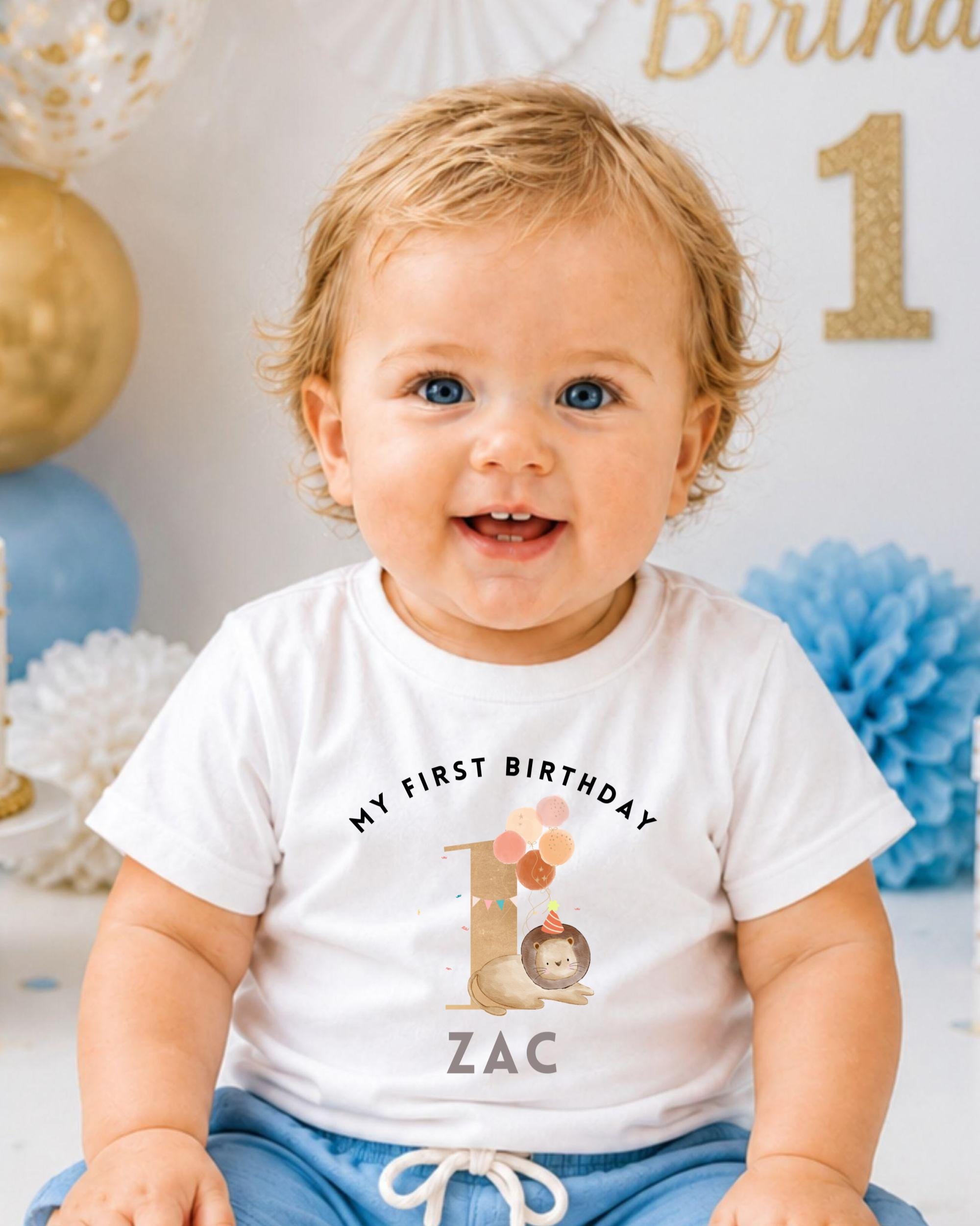 A smiling baby boy wearing a white t-shirt with a birthday message and a cartoon character on it, sitting in front of a birthday party decoration.