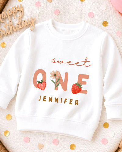 A white sweatshirt with a personalized text and graphics design, including the text "Sweet One" and a strawberry, along with the name "JENNIFER".