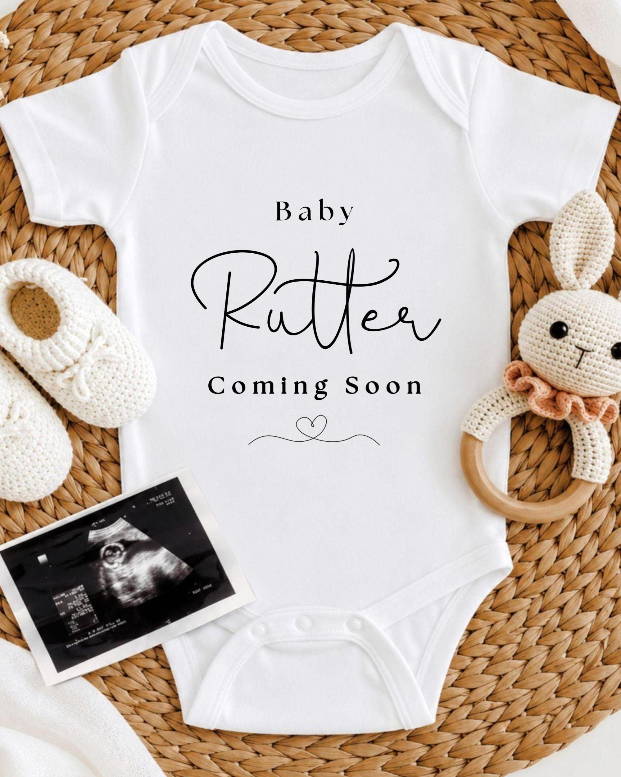 A white baby onesie with the text "Baby Rutter Coming Soon" printed on it, along with a crocheted rabbit toy and a black and white photograph of an infant's head.