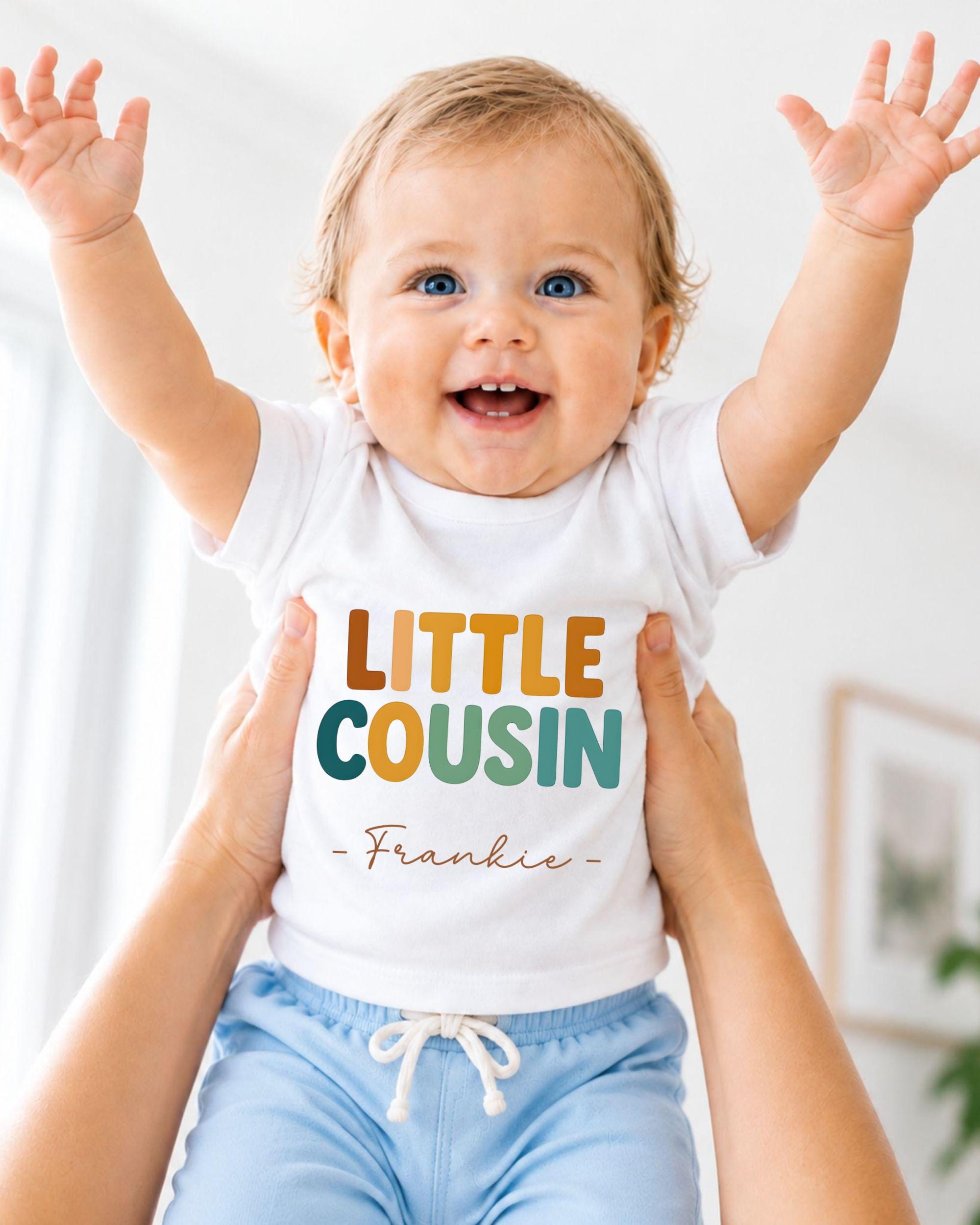 A smiling baby wearing a white shirt with the text "LITTLE COUSIN - Frankie" is being held up by someone's arms.
