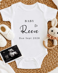 A white baby onesie with the text "BABY Reeve Due Sept 2028" printed on it, along with a crocheted bunny toy and a black and white photograph of an infant's head.