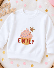 A white sweatshirt with a cupcake design and the name "EMILY" printed on it, surrounded by confetti.