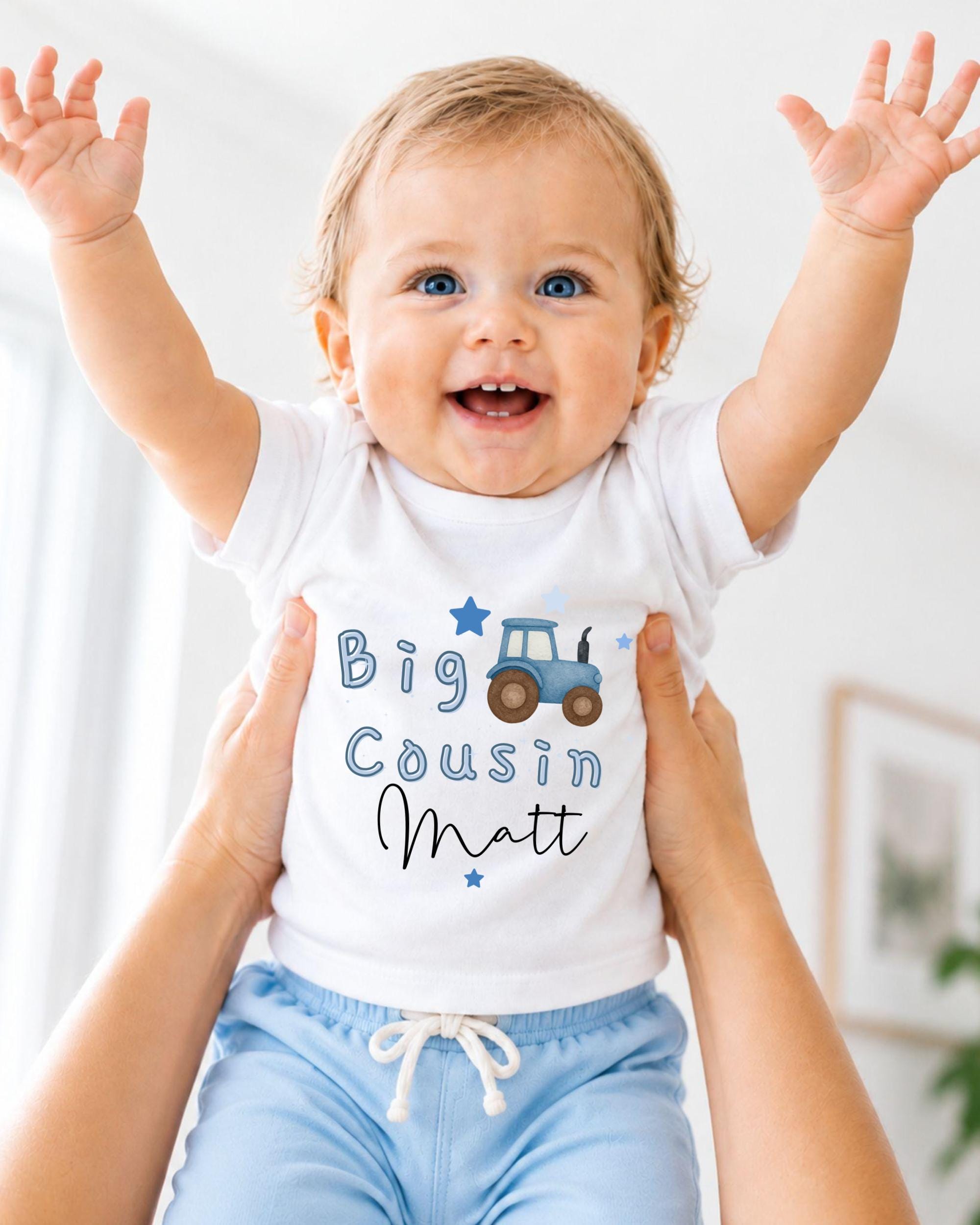 A smiling baby wearing a white shirt with a tractor graphic and the text "Big Cousin Matt" is being held up by someone's arms.