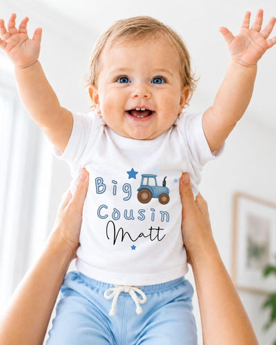 A smiling baby wearing a white shirt with a tractor graphic and the text "Big Cousin Matt" is being held up by someone's arms.
