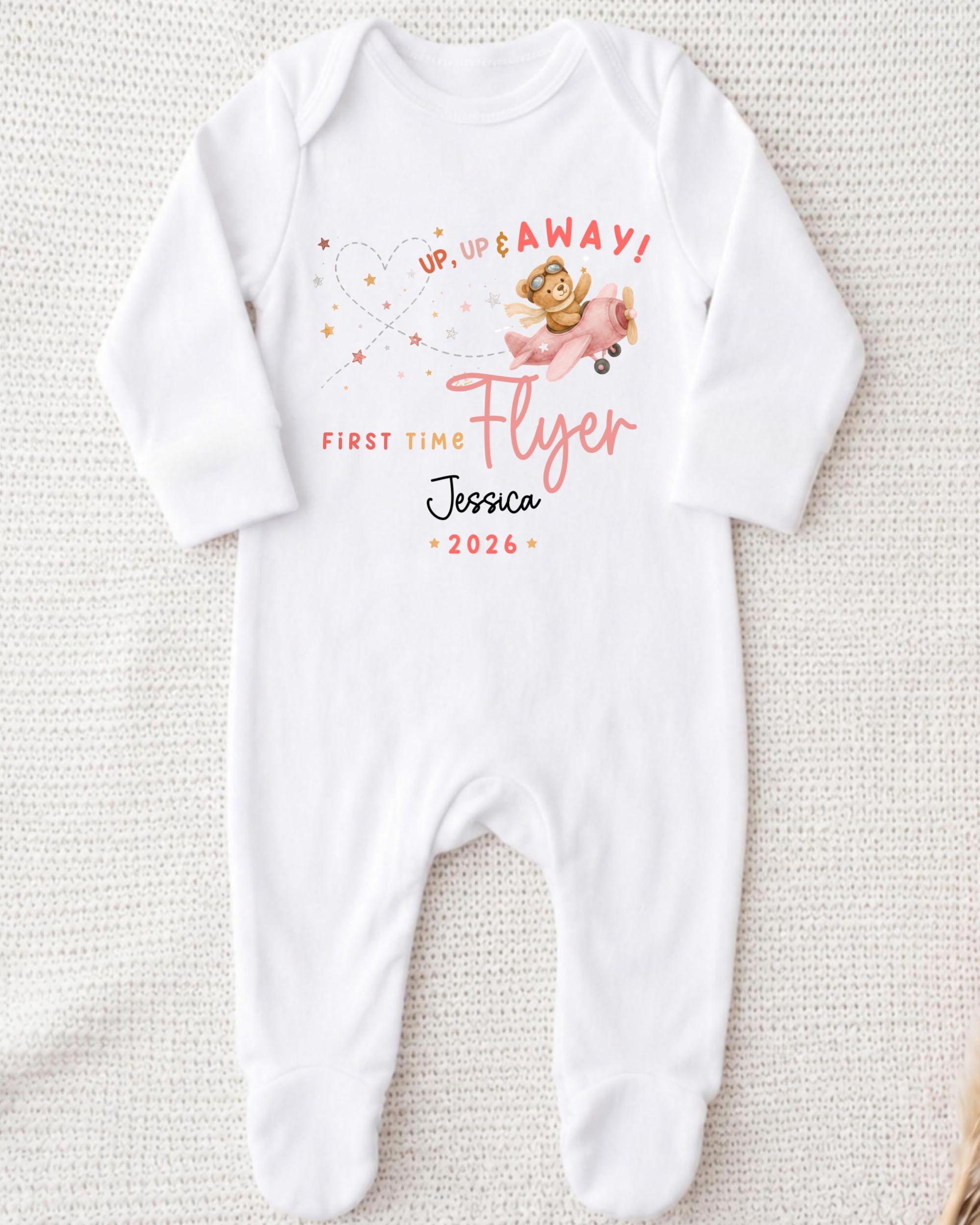 A white onesie with a printed design on it, featuring a teddy bear and the text "Up up away! First time flyer Jessica - 2026".