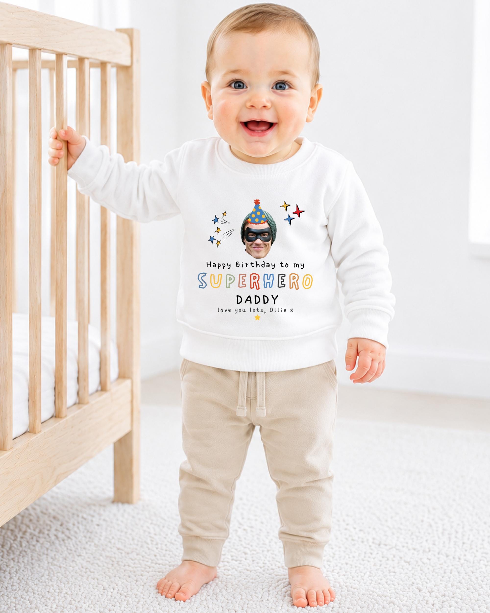 A smiling baby boy wearing a white sweatshirt with a superhero design and text, standing in front of a wooden crib.