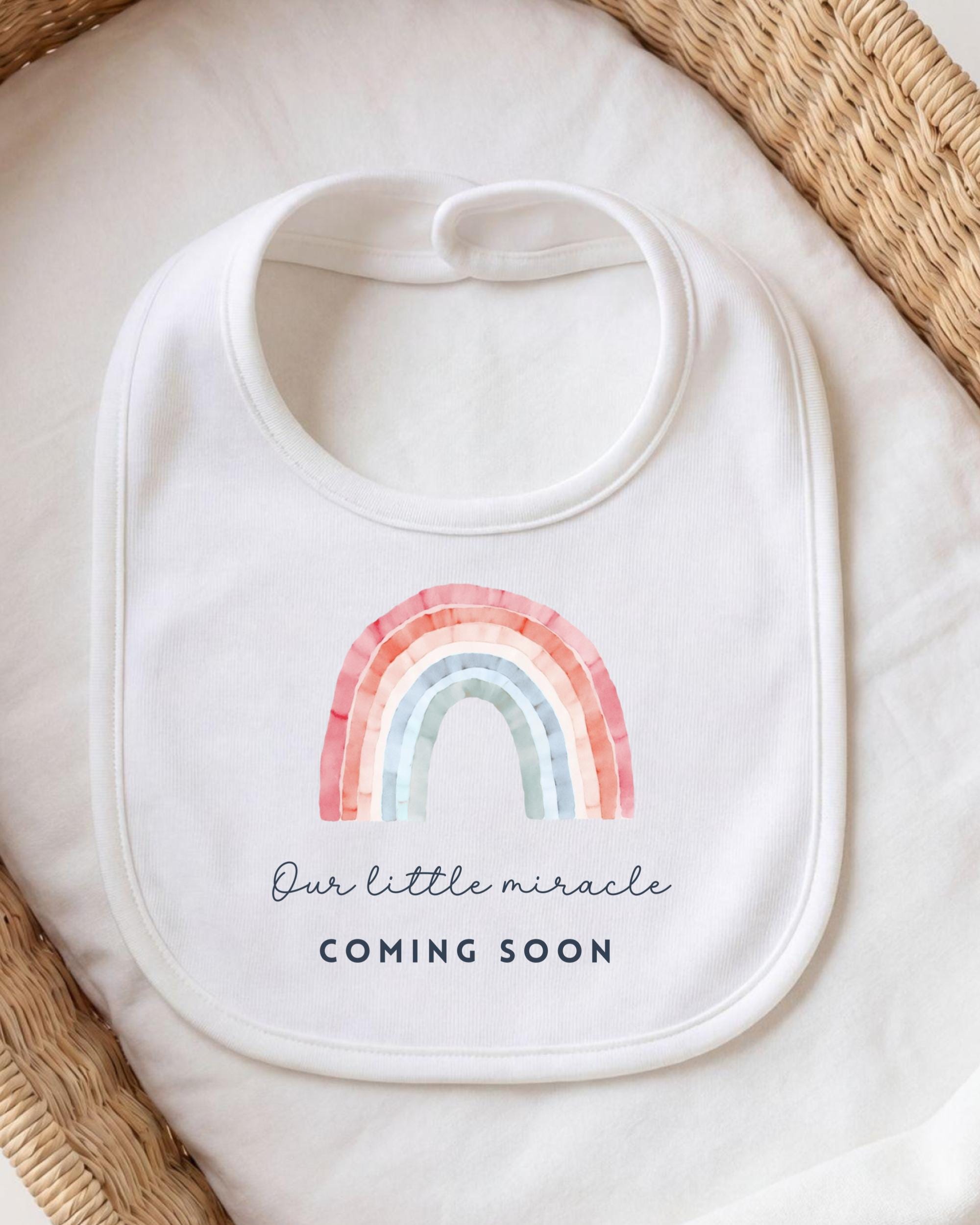 A white bib with a rainbow graphic and the text "Our little miracle coming soon" printed on it.
