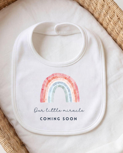 A white bib with a rainbow graphic and the text "Our little miracle coming soon" printed on it.