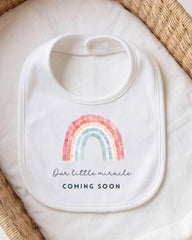 A white bib with a rainbow graphic and the text "Our little miracle coming soon" printed on it.