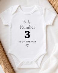A white baby onesie with the text "Baby Number 3 is on the way" printed on it.