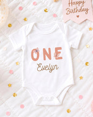 A white onesie with the text "ONE Evelyn" printed on it, surrounded by confetti and a birthday card.