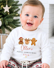 A smiling baby wearing a white sweater with a personalized Christmas-themed design, including a teddy bear, gingerbread man, and reindeer.