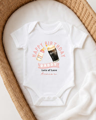A white onesie with a graphic design on it, featuring a bottle of milk and a glass of beer, along with the text "Happy Birthday Daddy Lots of Love Annie".