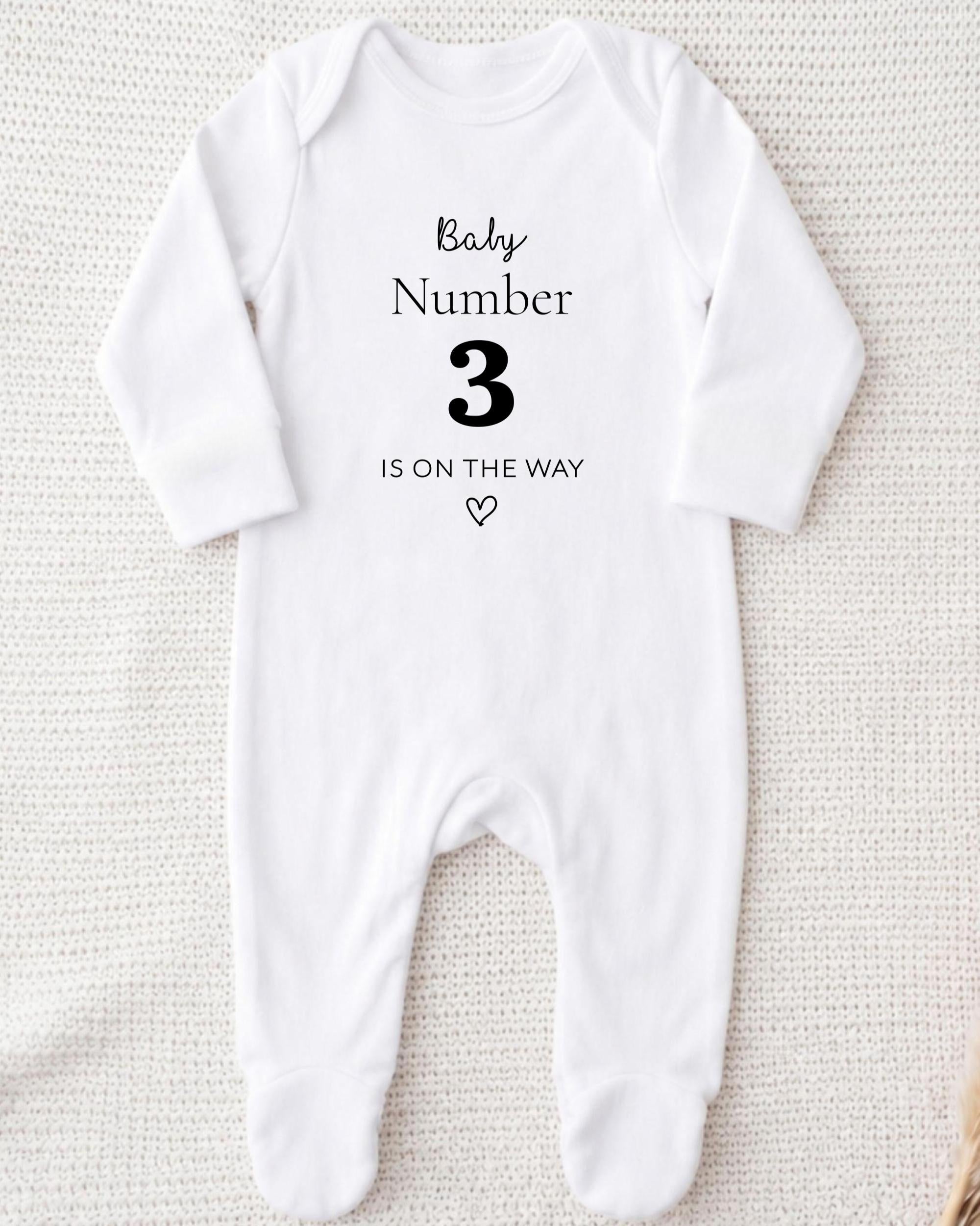 A white onesie with the text "Baby Number 3 is on the way" printed on it.