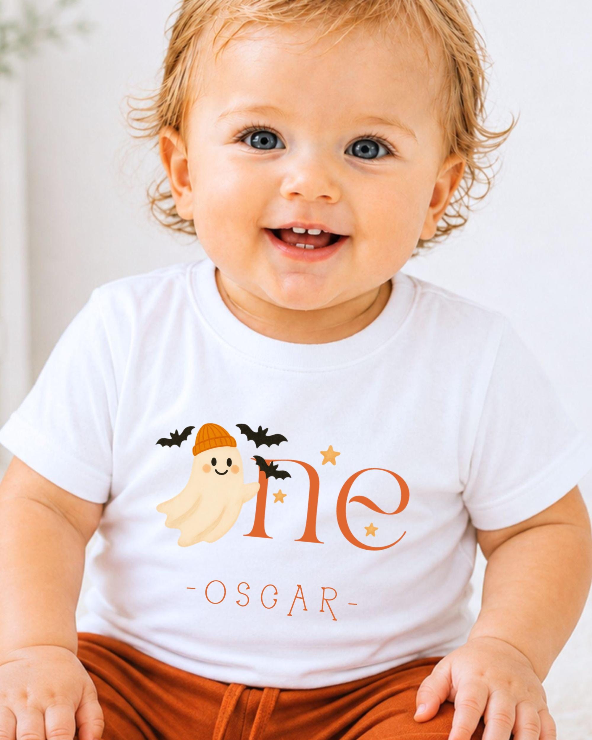 A smiling baby boy wearing a white t-shirt with a ghost and stars printed on it.