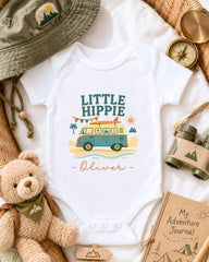 A baby onesie with a hippie-themed design, a teddy bear, a compass, a hat, and a book are arranged on a bed.
