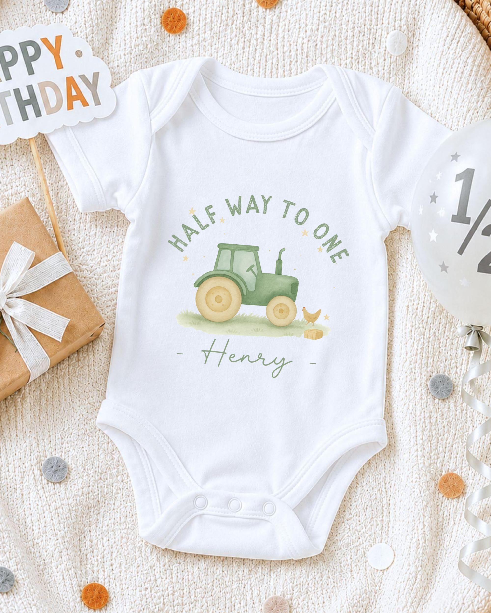 A white onesie with a tractor graphic and the text "Half Way to One Henry" printed on it, along with a birthday sign and confetti.