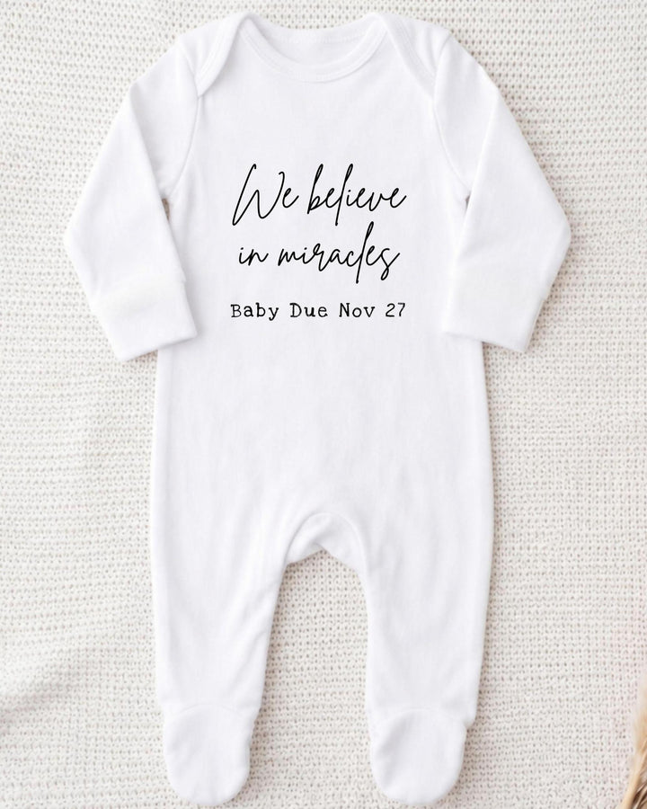 A white bodysuit with the text "We believe in miracles" printed on it, along with the date "Baby Due Nov 27".