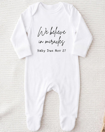 A white bodysuit with the text "We believe in miracles" printed on it, along with the date "Baby Due Nov 27".