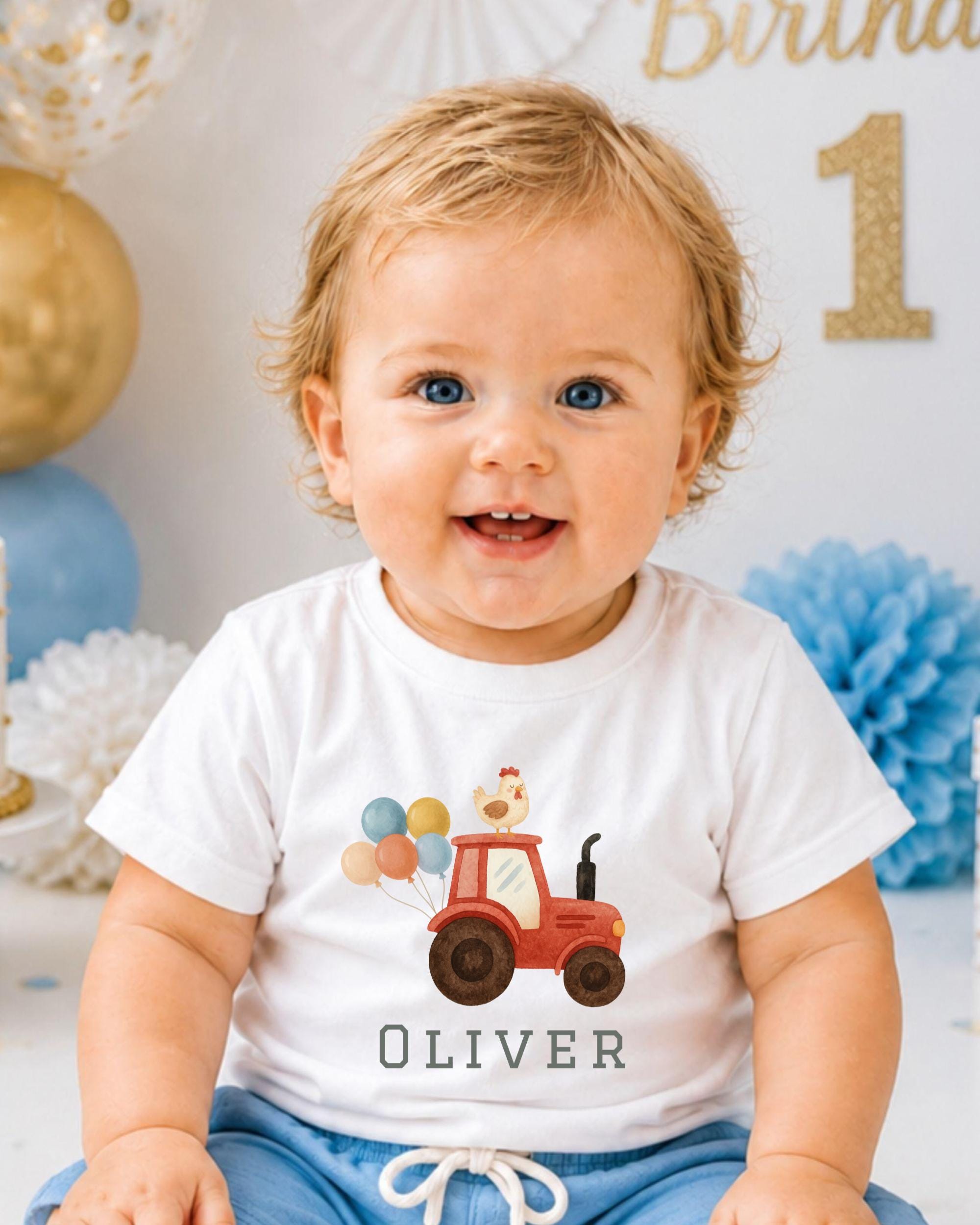 A smiling baby boy wearing a white t-shirt with a tractor and balloons printed on it, sitting in front of a birthday party backdrop.