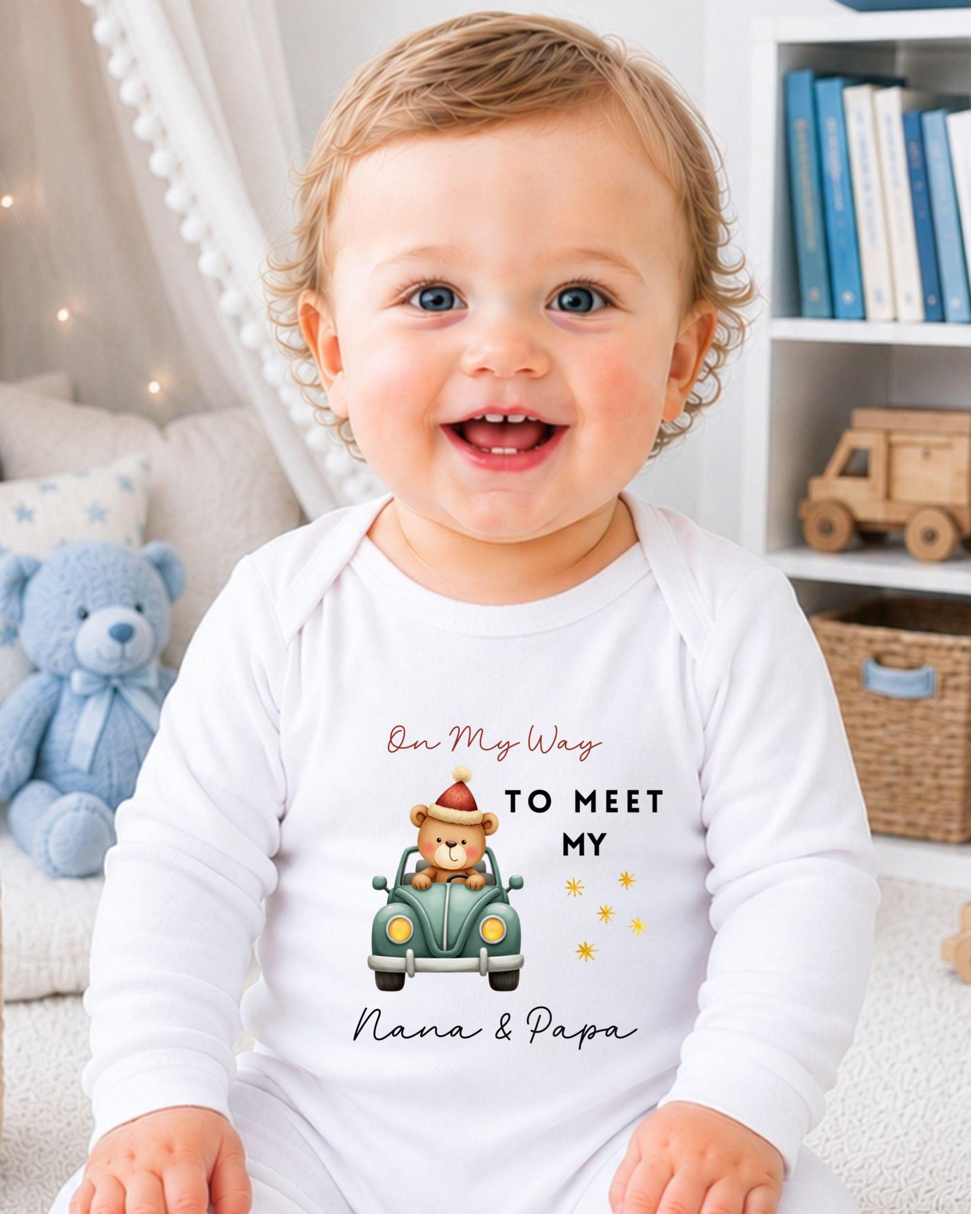 A smiling baby wearing a white onesie with a cartoon image of a bear driving a car and text that says "On My Way to Meet My Nana & Papa".
