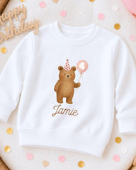 A white sweatshirt with a cartoon bear wearing a party hat and holding a balloon, along with the name "Jame" printed on it.