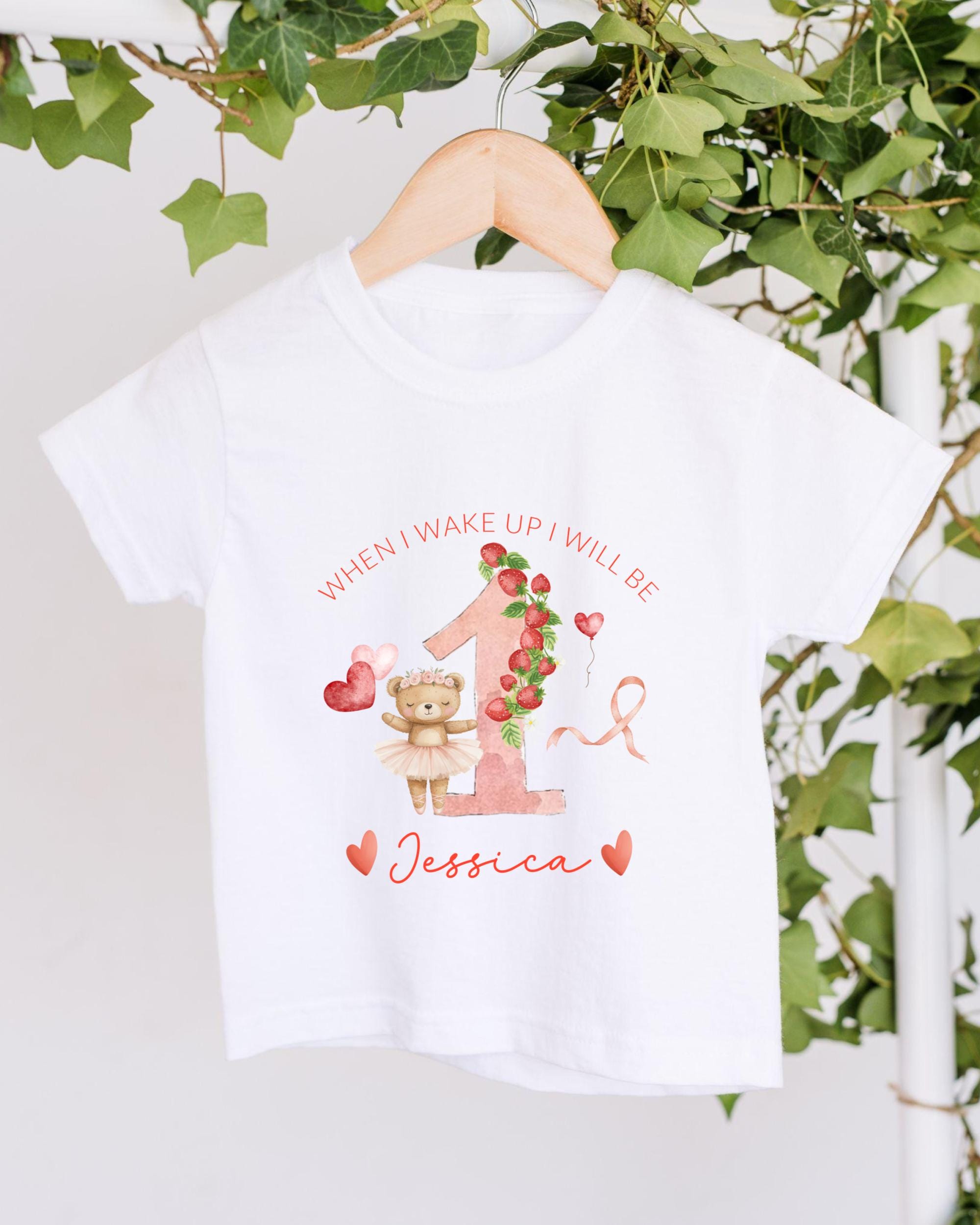 A white t-shirt with a personalized design hanging on a wooden hanger. The design features a teddy bear, a pink number one, and the text "When I wake up I will be Jessica".