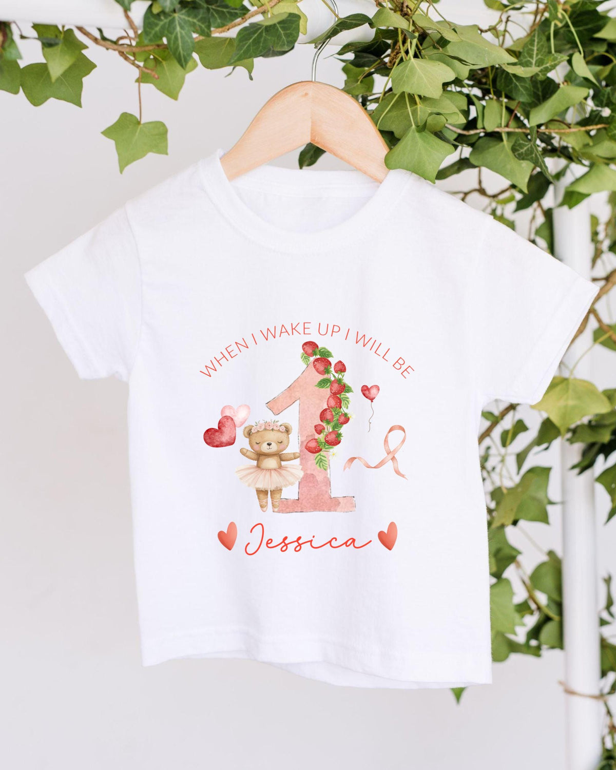 A white t-shirt with a personalized design hanging on a wooden hanger. The design features a teddy bear, a pink number one, and the text "When I wake up I will be Jessica".