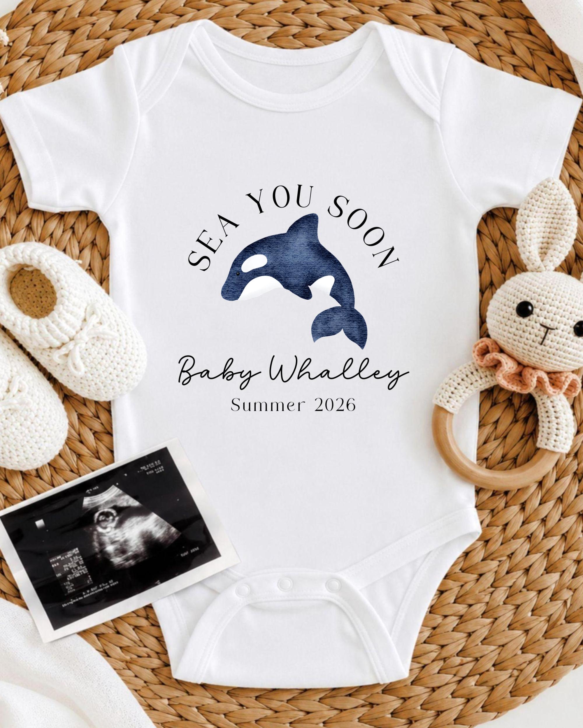A white onesie with a graphic of a whale and the text "SEA YOU SOON Baby Whalley Summer 2026" is displayed on a woven mat, accompanied by a crocheted bunny toy and a baby's first photograph.
