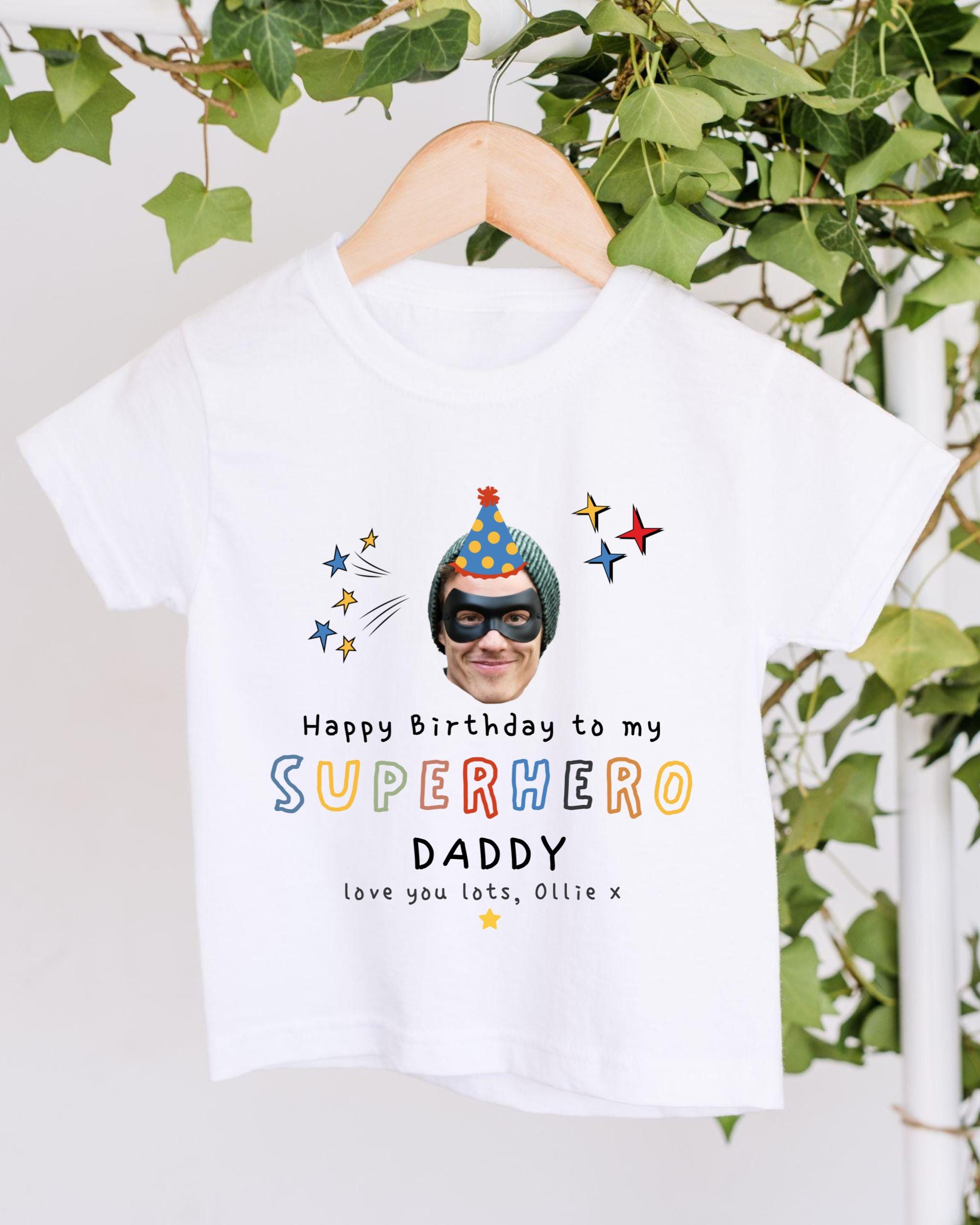 A white t-shirt with a printed design featuring a superhero character and text that reads "Happy Birthday to my SUPERHERO DADDY love you lots, Olilee".
